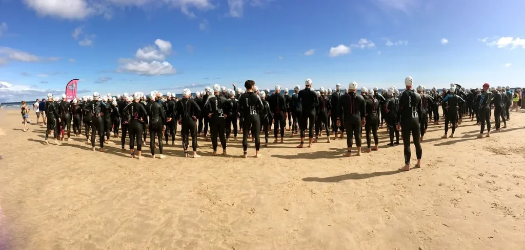 Zawodnicy w piankach i czepkach zebrani na plaży przed startem pływania w triathlonie